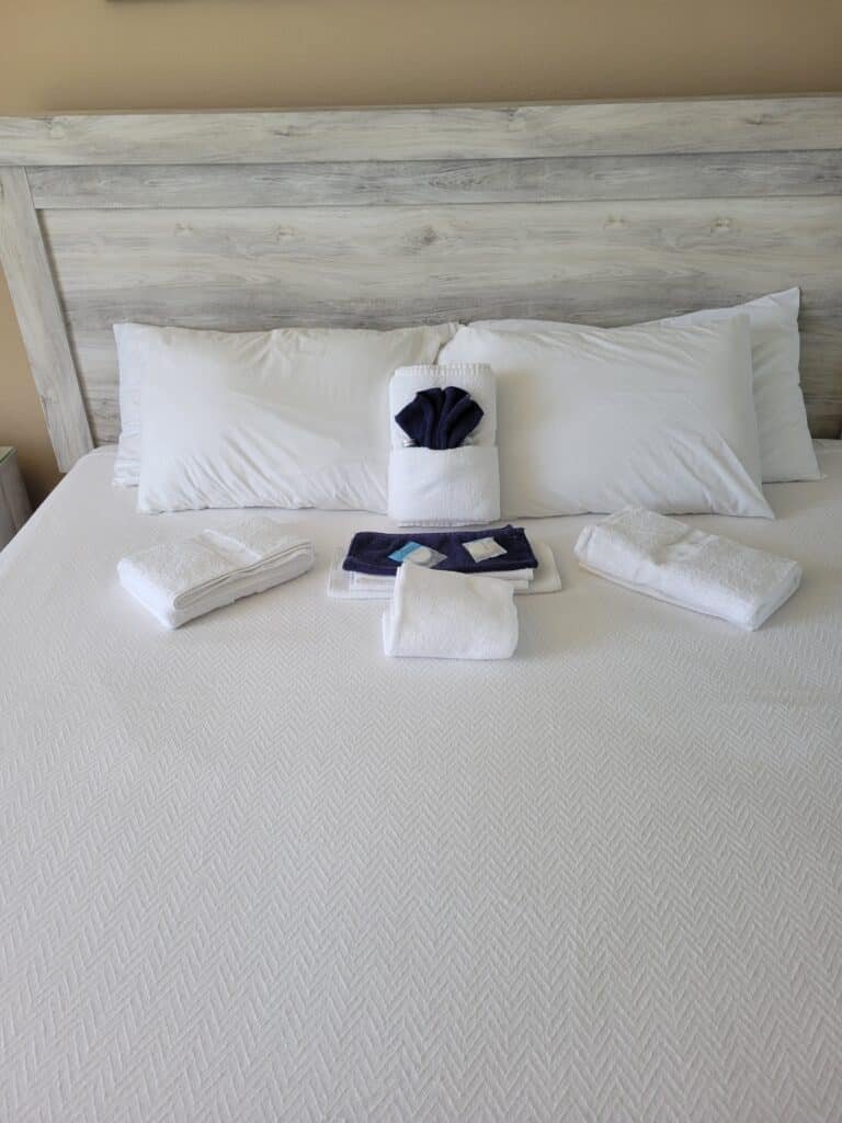 20230607_140334 - Mister Moppin Freshly made hotel-style bed with a wood headboard, folded white bath towels, and a navy welcome towel set staged after a post-clean inspection